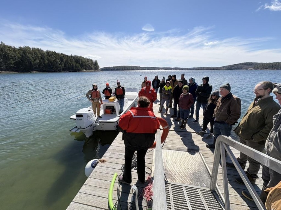 A group of people listens to an instructor on a dock beside a small motorboat on a sunny day by a wooded shoreline.