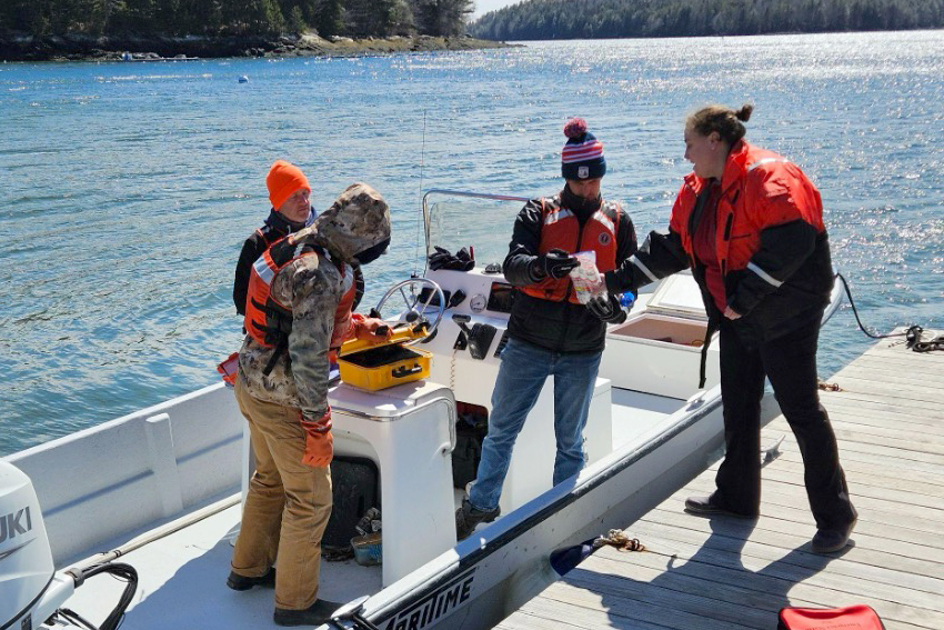 Three people in life jackets stand near a small boat docked at a wooden pier, with water and trees visible in the background.
