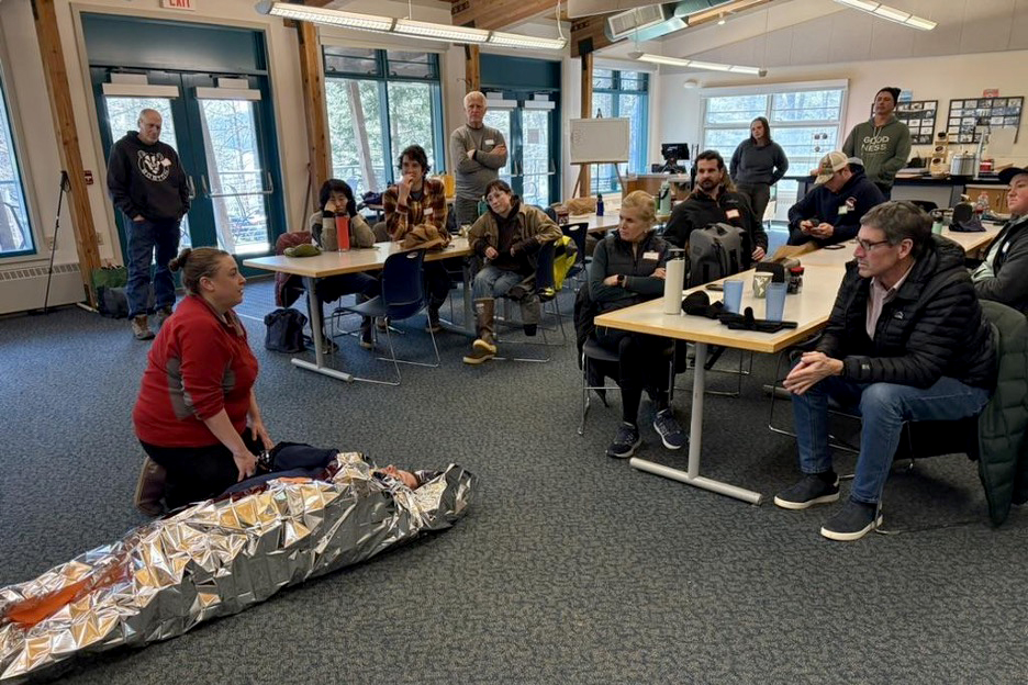 A group of adults watches as an instructor kneels next to a person wrapped in a thermal emergency blanket during an indoor demonstration.