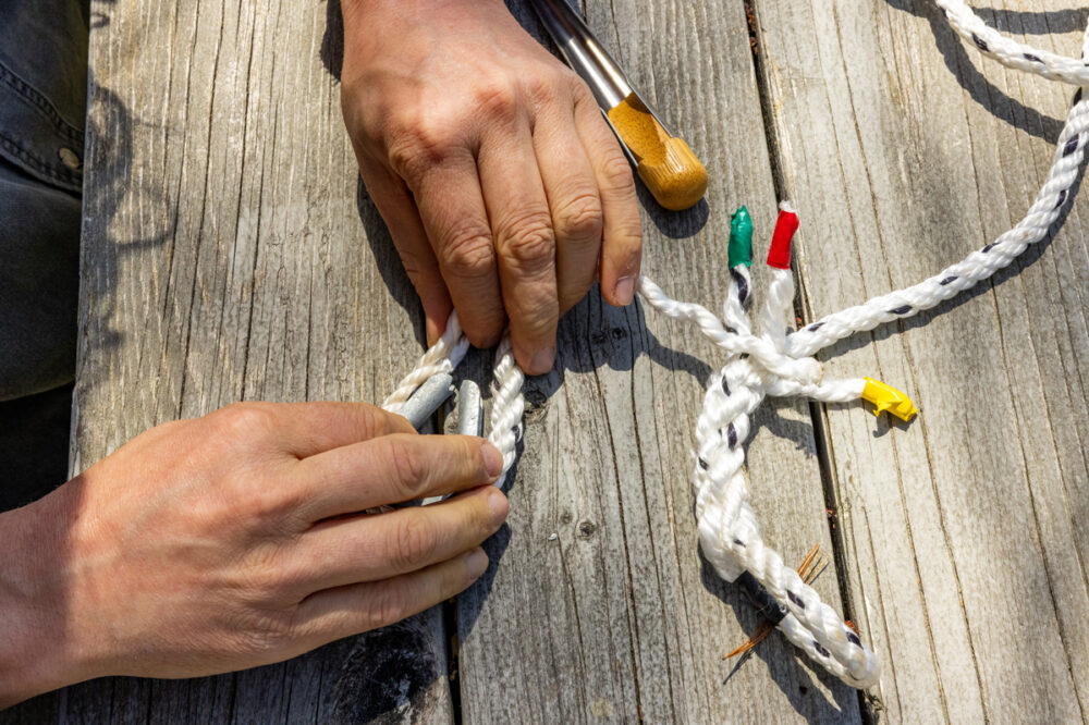 Two hands are splicing a white rope on a wooden surface, with three rope ends marked by green, red, and yellow tape nearby.