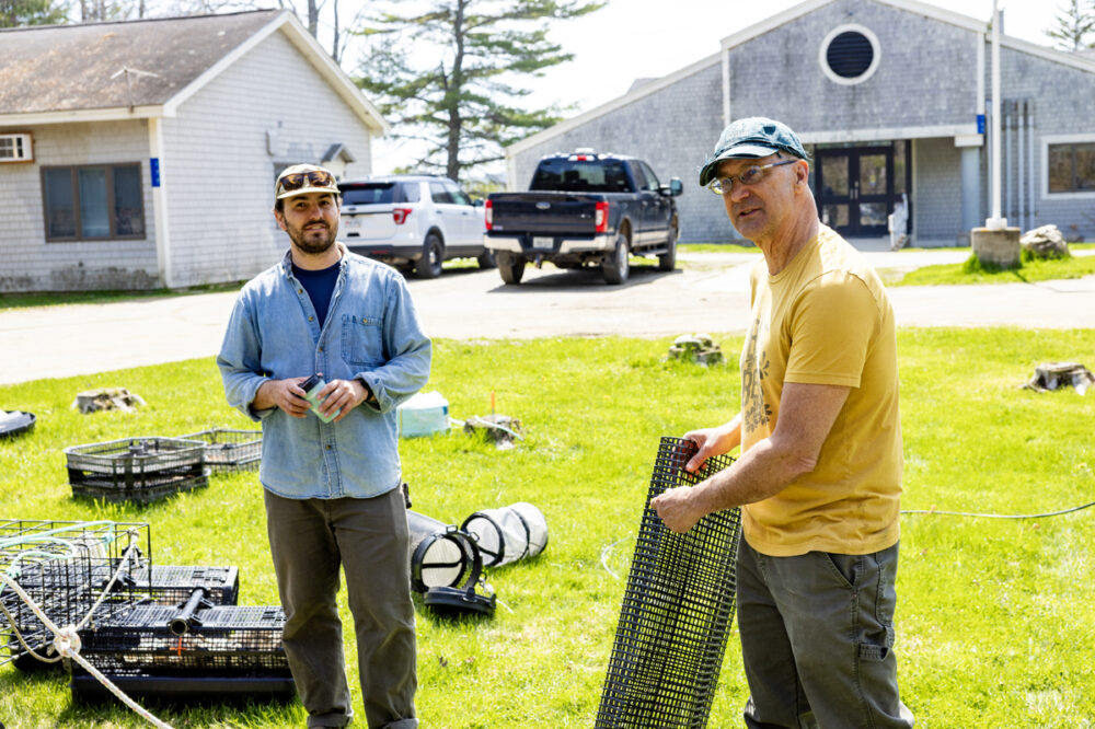 Two men stand on grass with crab traps and fishing equipment, near a building and parked trucks, on a sunny day.