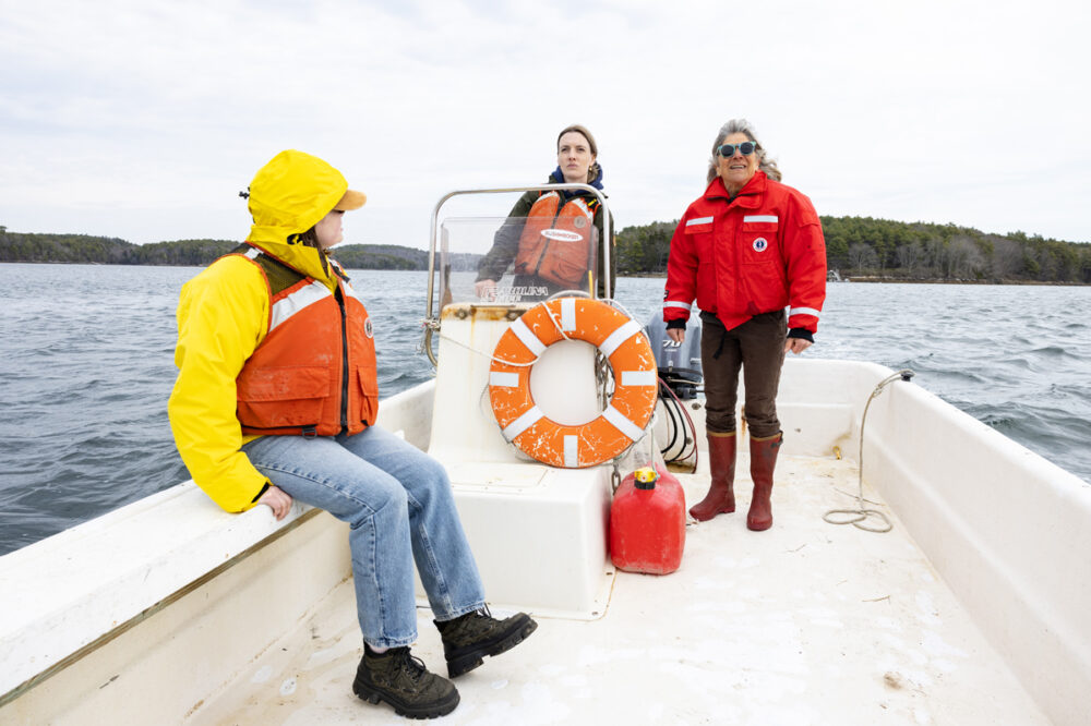 Three people in jackets and life vests are on a small boat on the water, with trees visible in the background and a life ring attached to the console.