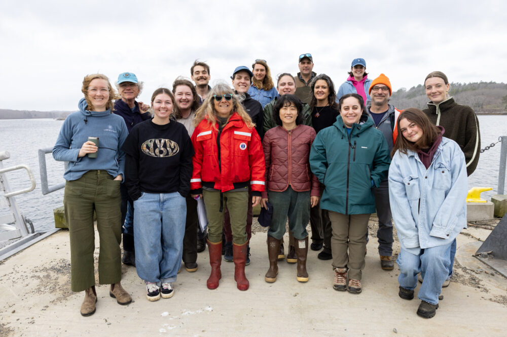 A group of eighteen people stand and smile together outdoors on a dock by the water, dressed in casual and outdoor clothing, with a cloudy sky in the background.