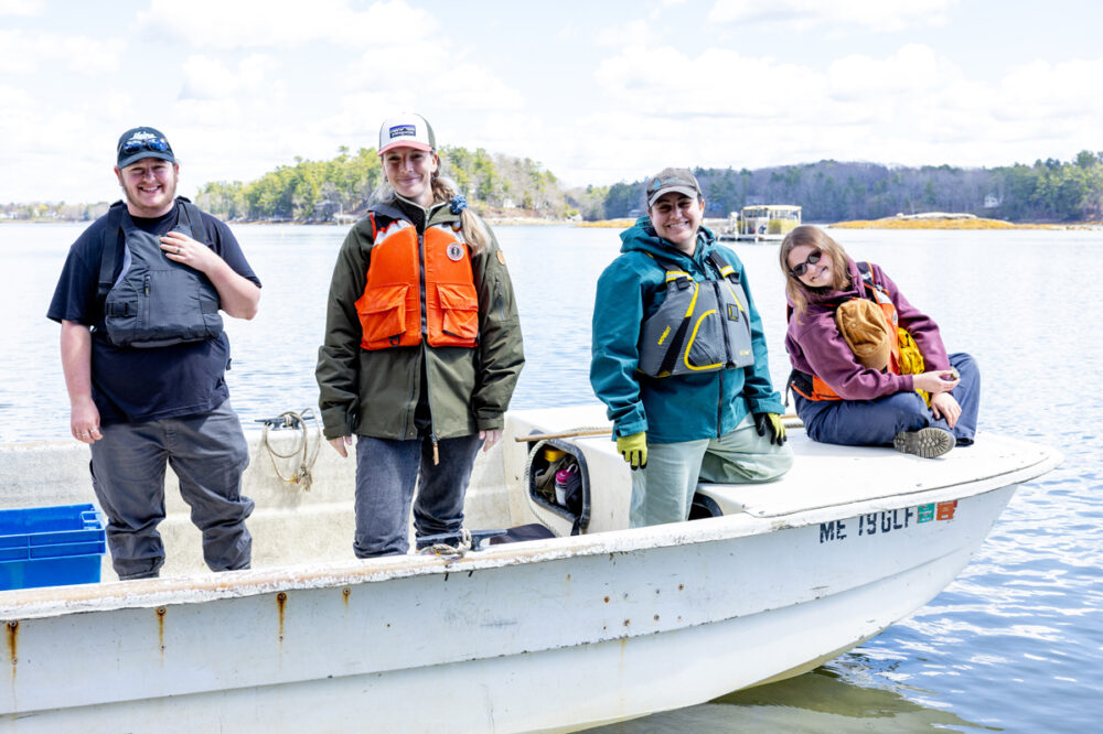 Four people wearing life jackets stand and sit on a small boat floating on the water, with trees and buildings visible in the background.