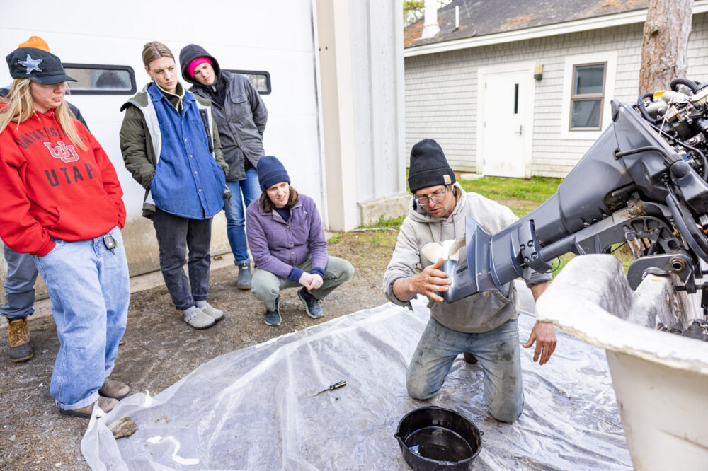 A person kneels on a tarp while repairing a boat motor, with four people standing and watching attentively outside a building.
