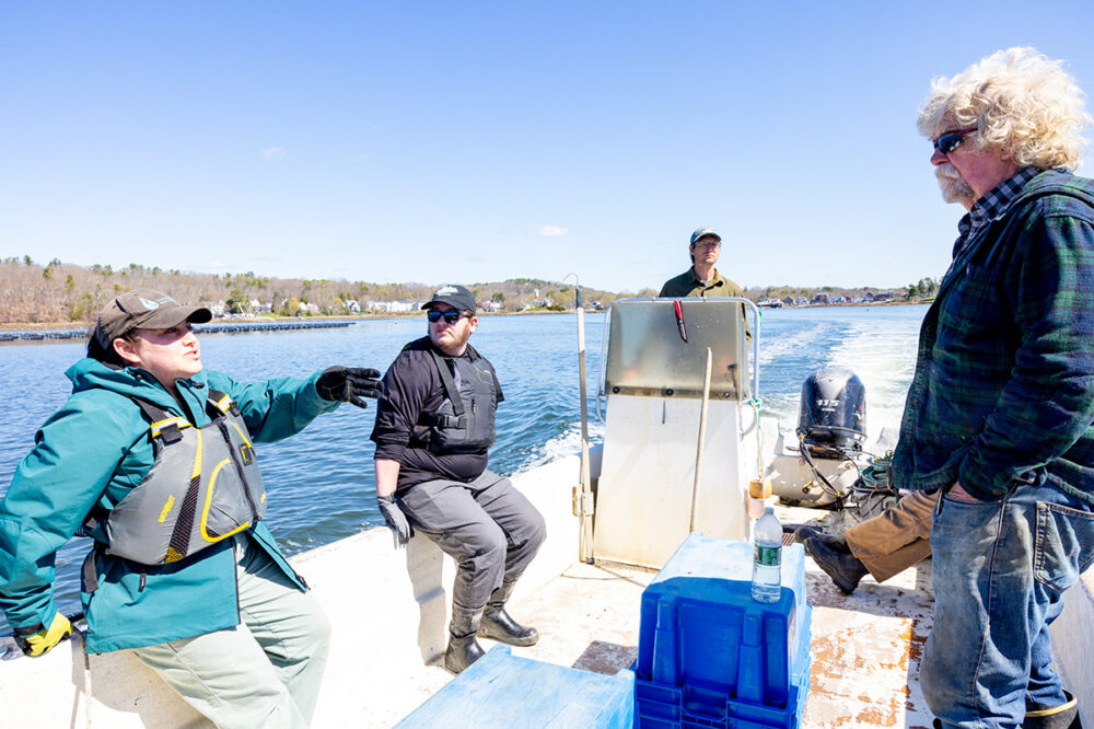 Four people ride in a small motorboat on a sunny day, with one person speaking and others listening; trees and houses are visible along the shoreline in the background.