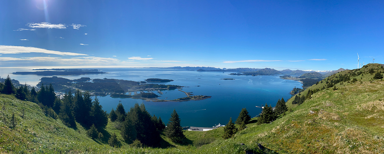 Panoramic view from a grassy hillside overlooking a bay with islands, distant mountains, scattered clouds, and a clear blue sky.